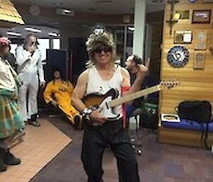 A man in an 80s wig holding an electric guitar and making the 'horn' sign