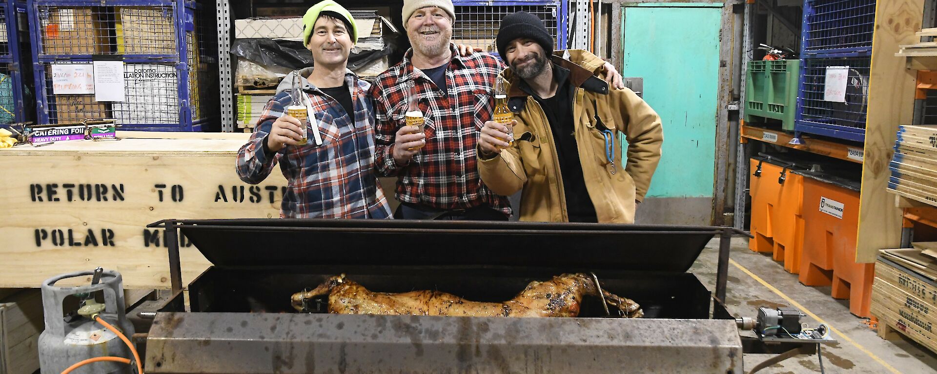 Three expeditioners standing behind the lamb spit, shoulder to shoulder with beers in hand and big grins on their faces.