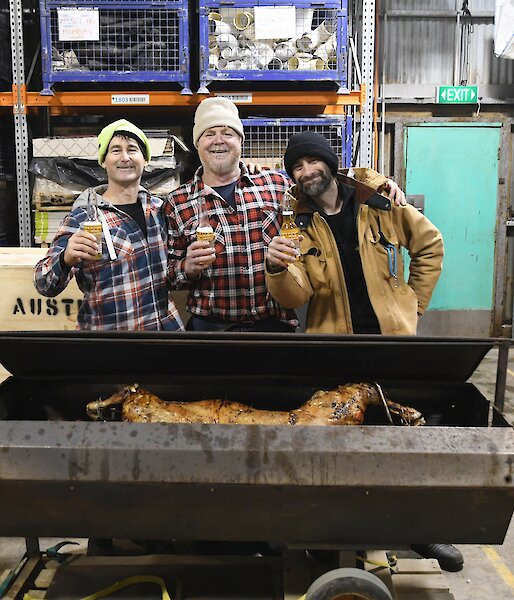 Three expeditioners standing behind the lamb spit, shoulder to shoulder with beers in hand and big grins on their faces.