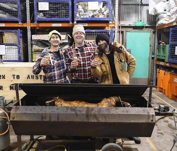 Three expeditioners standing behind the lamb spit, shoulder to shoulder with beers in hand and big grins on their faces.