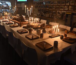 A long table with white chairs and tablecloth is set with candelabras, handmade wooden boards for plates, rustic handmade forks and handmade wooden mugs with copper bands around their base.  The food is laid out, with plates of bread, bowls of vegetables, plates of chicken, and a whole lamb on a large wooden carving board is in the centre.