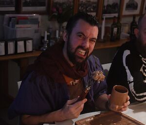 Jamie sitting at the table, wearing a blue robe, a brown scarf, a black beard and slicked back hair, and a snarl on his face. In one hand is as wooden mug, and in the other a fork with a large chunk of meat.