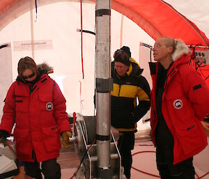 Three scientists standing beside an ice core drill in a tent in Antarctica.