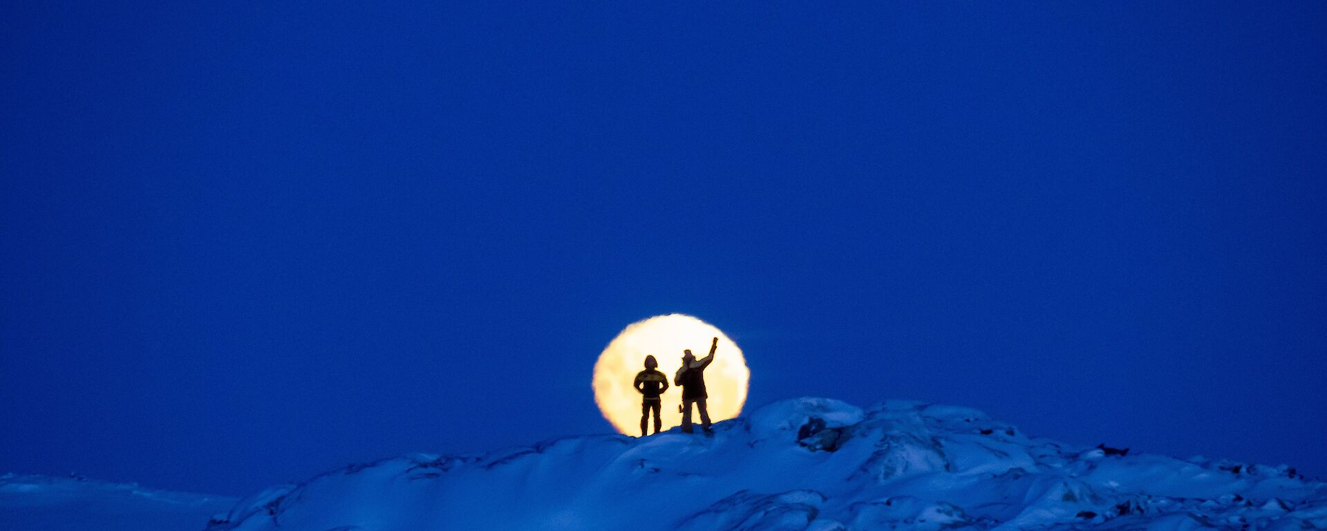 Two expeditioners silhouetted against the moon whilst standing on a snowy mountain