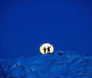 Two expeditioners silhouetted against the moon whilst standing on a snowy mountain