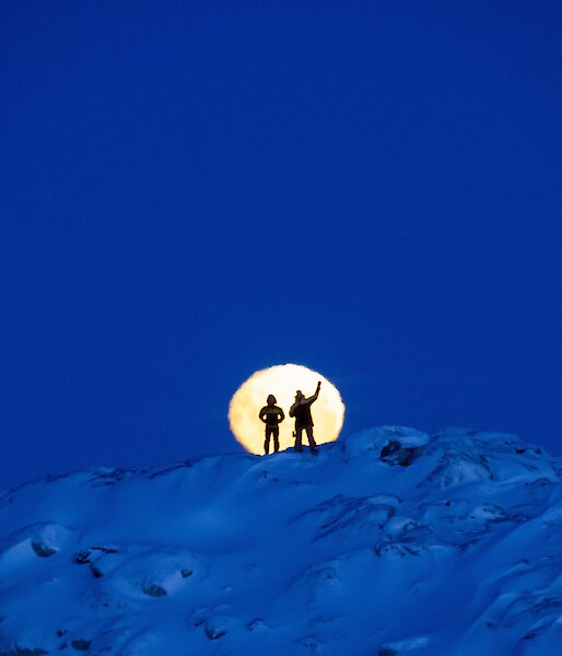 Two expeditioners silhouetted against the moon whilst standing on a snowy mountain
