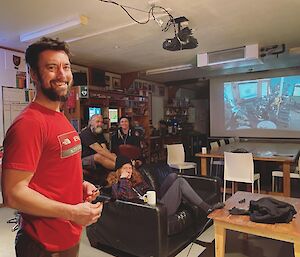 A player posing with darts in his hand. The team can be seen in the background watching the darts game on the big screen.