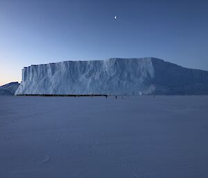 Night shot of an iceberg in distance with the moon above and penguins at the base