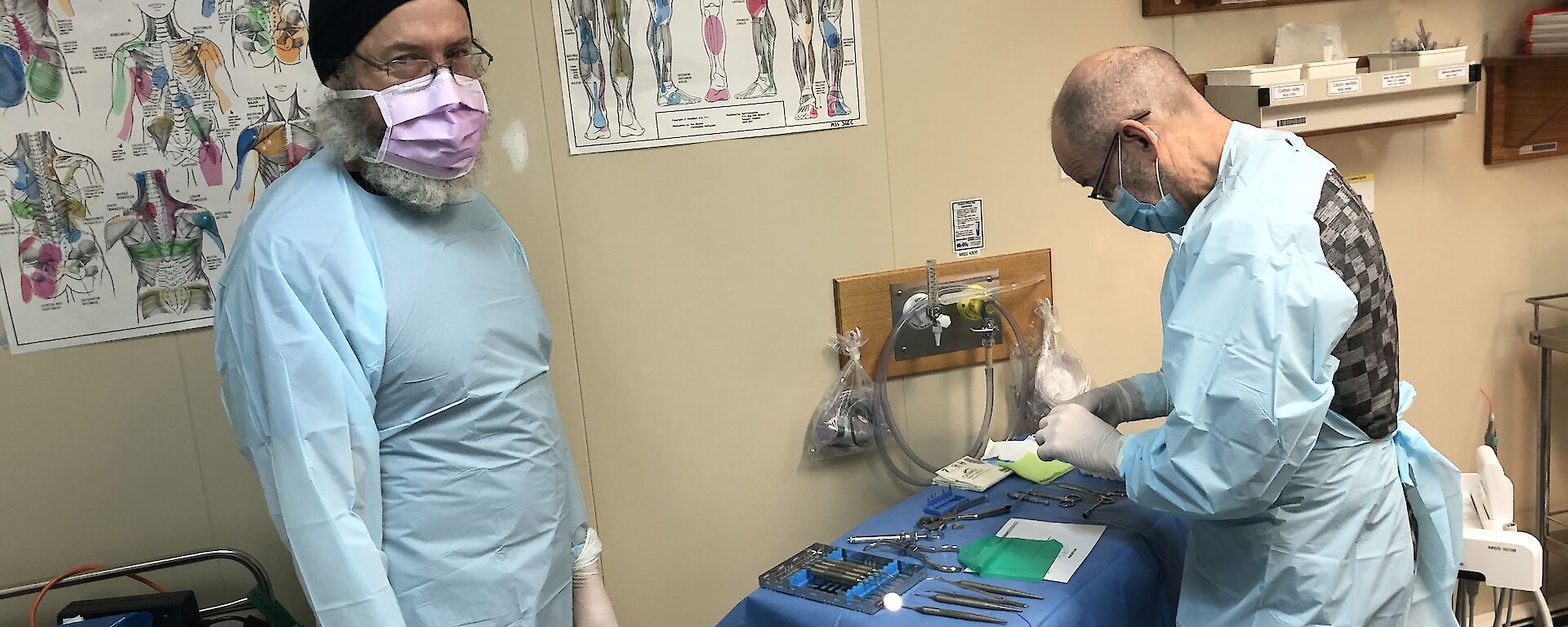 Two men in scrubs and face masks prepare the dentistry equipment in the medical room