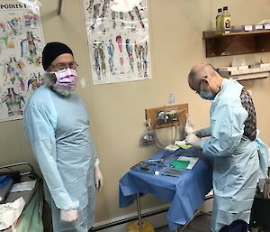 Two men in scrubs and face masks prepare the dentistry equipment in the medical room