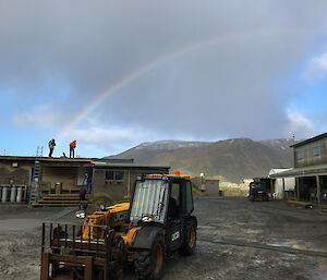 Two workers on the roof silhouetted against a blue cloudy sky with a mountain in background complete with a rainbow.  Tractor in foreground.