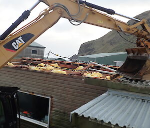 Yellow arm of an excavator reaches up over the top of a building with it's bucket resting on the roof sheets to hold them down.