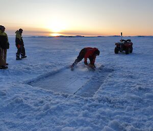 Snowy landscape with an expeditioner cutting a rectangle into the ice to create a sea ice pool.  Sunset in the background as colleagues look on.