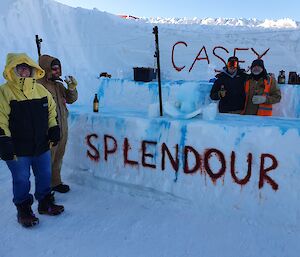 A bar made out of ice with 'Splendour' written in red in to the ice.  Two men stand behind the bar serving with two expeditioners being served in the foreground.  Casey sign hanging in the background.