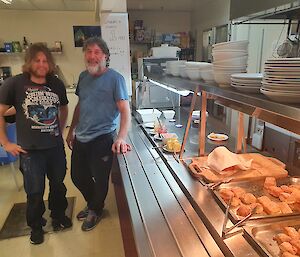 Two expeditioners in the kitchen smiling to camera with hot plates of food to their left