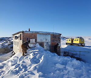The door of the Wilkes hut blocked with snow.  An expeditioner is shovelling the snow.  Yellow Hagglund in background.