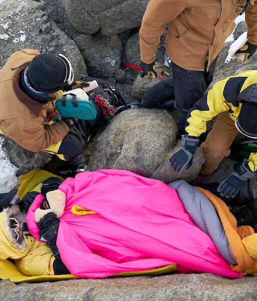 An aerial shot looking down at the injured expeditioner, lying on the ground covered in warm sleeping bags.  Various rescuers around him