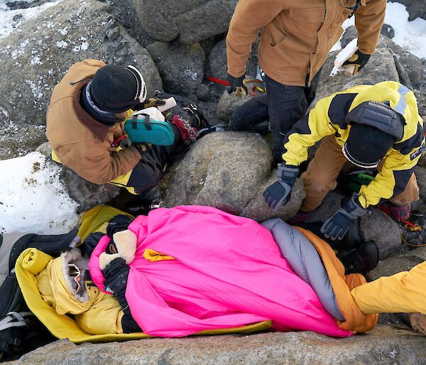 An aerial shot looking down at the injured expeditioner, lying on the ground covered in warm sleeping bags. Various rescuers around him