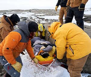 Four rescuers lift up the injured expeditioner on a stretcher while two others look on from the rocks