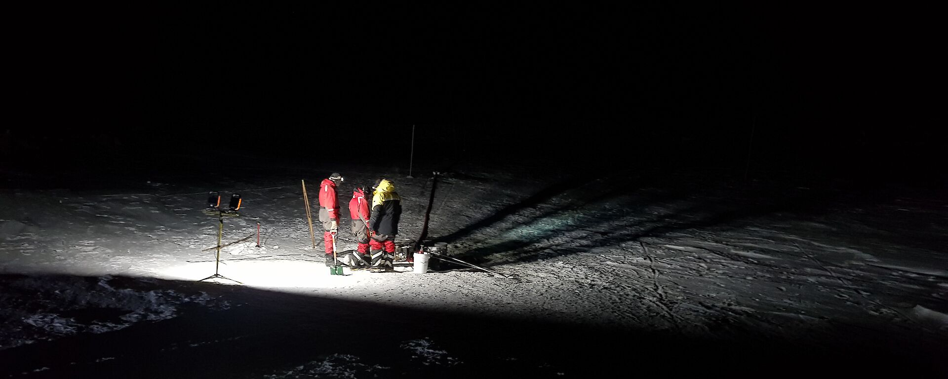 Pitch black night with three men working under lights in the centre of the picture