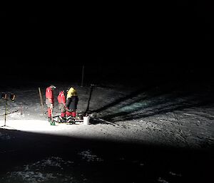 Pitch black night with three men working under lights in the centre of the picture