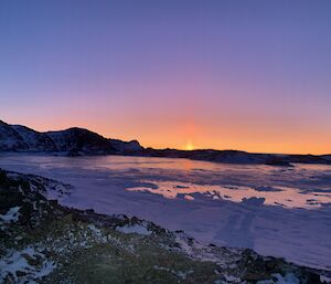 Rocky and snowy landscape with the sun coming over the mountains on the horizon creating a orange sunrise