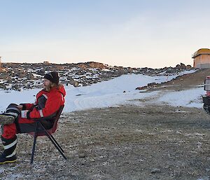 An expeditioner sitting on a chair, outside in the snow, looking at the view.  A Hagglund is parked behind him.