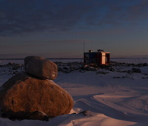 Jacks hut near Casey, lit by a low winter sun.  Two large rocks, one on top of the other, in foreground