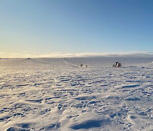 Blue skies and snow from foreground to horizon with a convoy of vehicles setting off for Wilkins from Casey making tracks in to the distance.