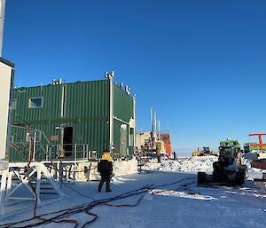 Clear blue sky with large green shed building at the centre, cables on the gound, expeditioners working and various tractors and lorries