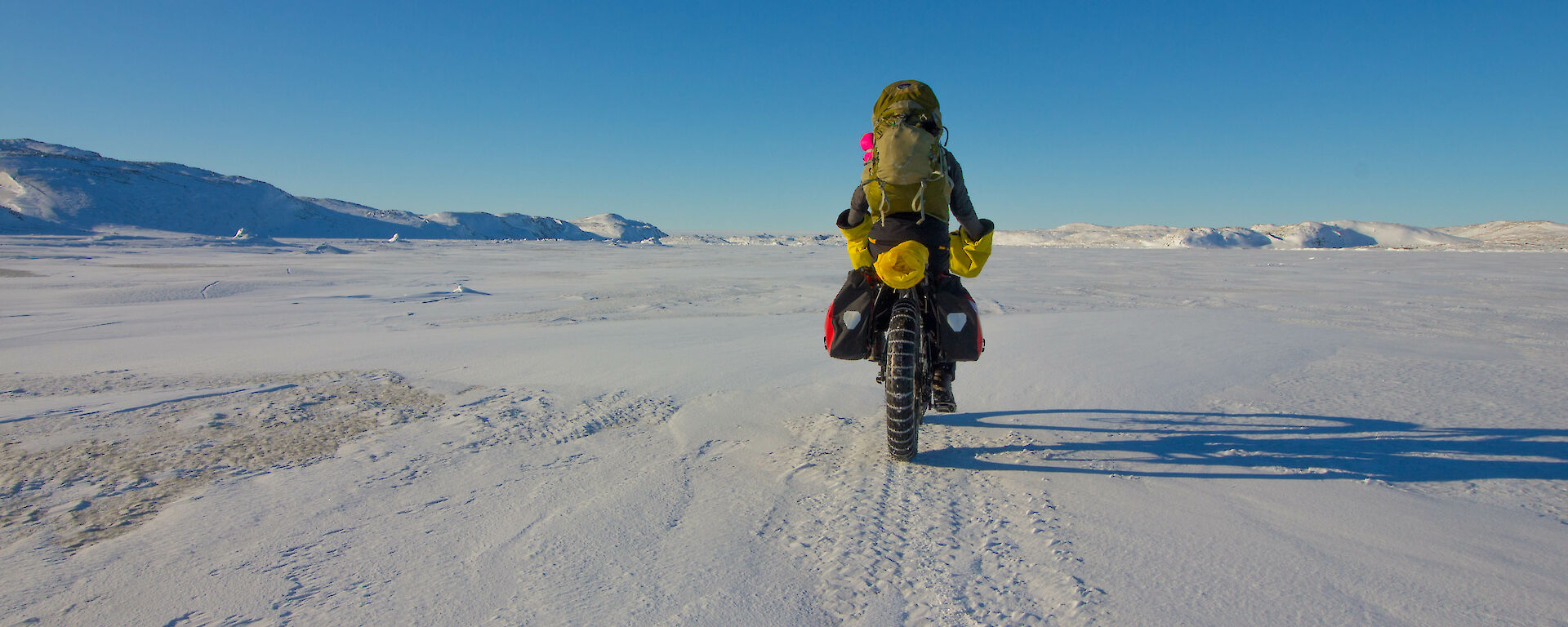A biker loaded with gear riding away from camera across the ice towards a beautiful blue skied horizon