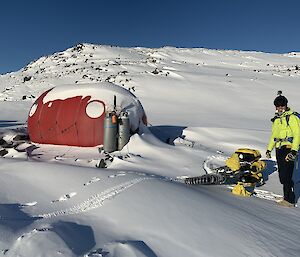 An expeditioner standing beside a red 'melon' (small caravan) half buried under some snow
