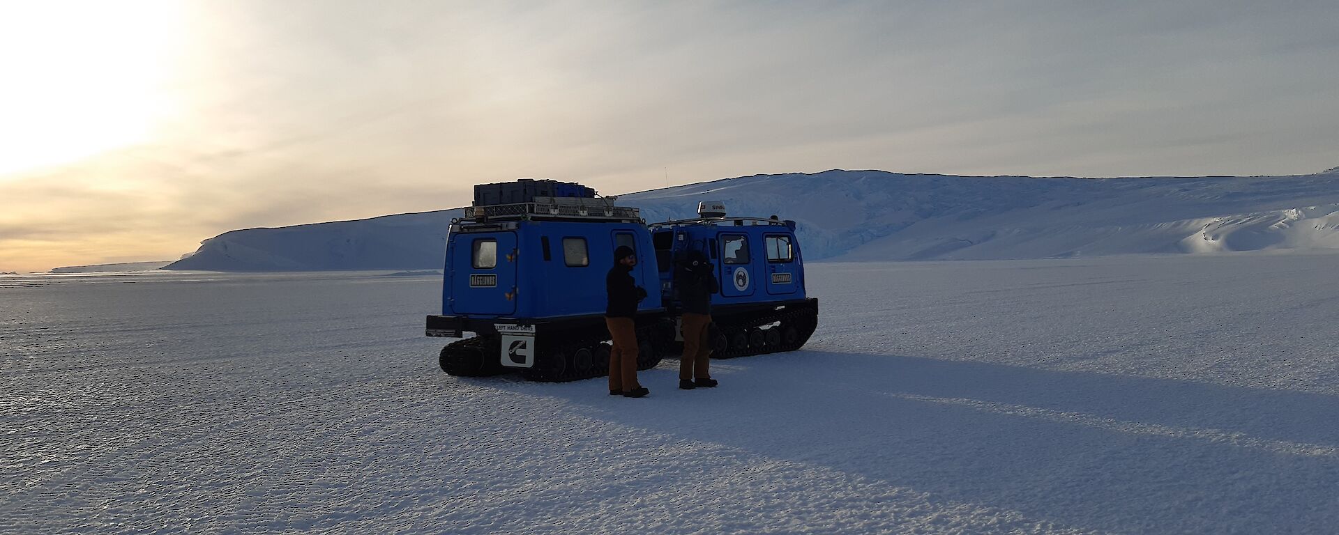 Two blue Hagglunds in the snow with two expeditioners in front