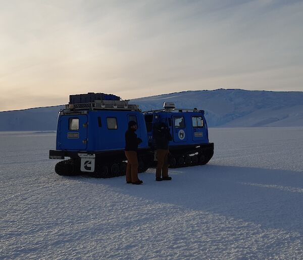 Two blue Hagglunds in the snow with two expeditioners in front