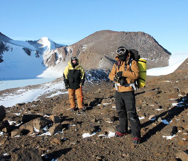 Two expeditioners in cold weather gear face camera with frozen lake in background