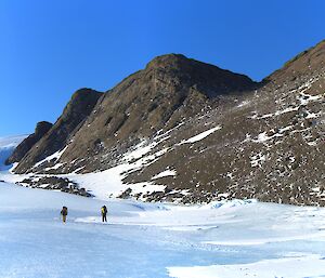 Two expeditioners walk off in the distance across a frozen lake with rocky hill on their left