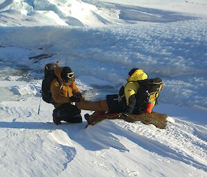 Two expeditioners in an icy landscape.  One expeditioner kneels and rubs the others foot.