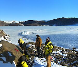Three expeditioners take a break with backpacks on ground with frozen Lake Henderson behind them