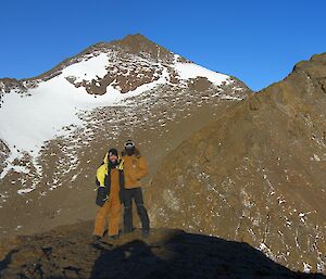 Two expeditioners stand on the top of the mountain with large moutains in background