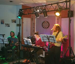 Three expeditioners in a band playing instruments with music stands, microphones and lights