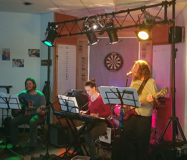 Three expeditioners in a band playing instruments with music stands, microphones and lights