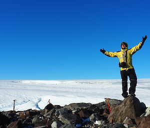 A man on top a hill with his arms out spread and a snow and clear blue skies in the background