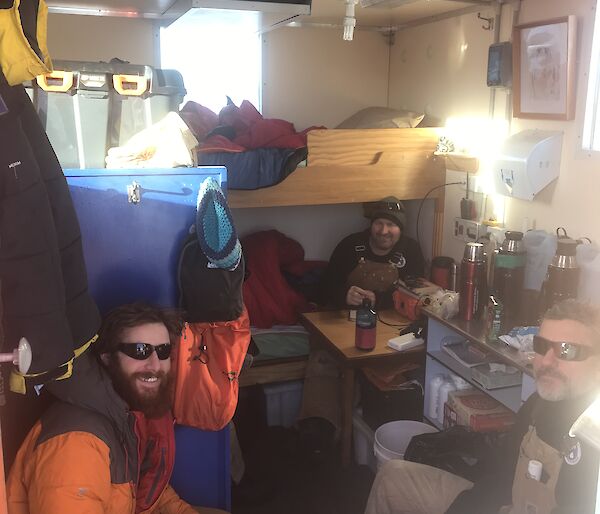 Four expeditioners inside a small hut sitting eating. Bunk style beds in background.