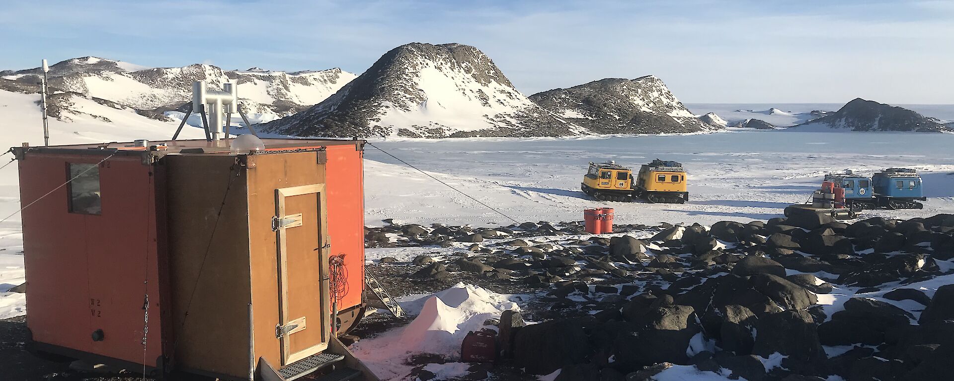 A small red hut in the foreground with two Hägglunds behind.  Ice covered hill in the distance.
