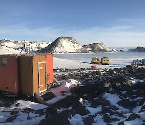 A small red hut in the foreground with two Hägglunds behind.  Ice covered hill in the distance.