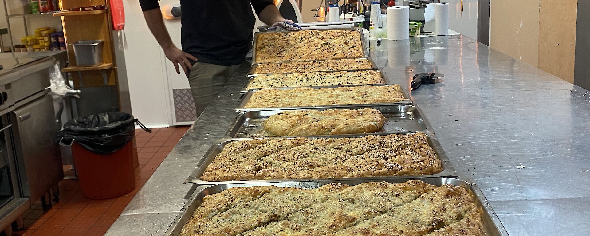 7 trays of fresh made borek on a kitchen bench with an expeditioner smiling to camera