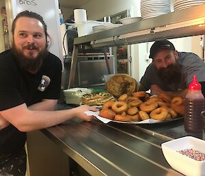 Two expeditioners at the station kitchen pass with a large platter of fresh made doughnuts