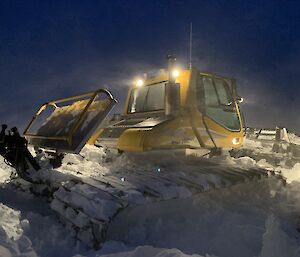 A snow and ice bound groomer half dug out at night with its lights on