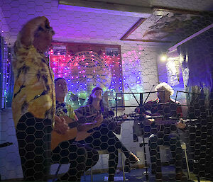 Man in Hawaiian shirt standing in front of three people sitting playing acoustic guitar