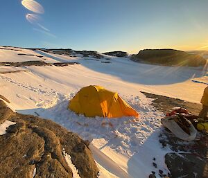 A yellow tent set up in the snow with rocks around.  An expeditioner stands to the side.